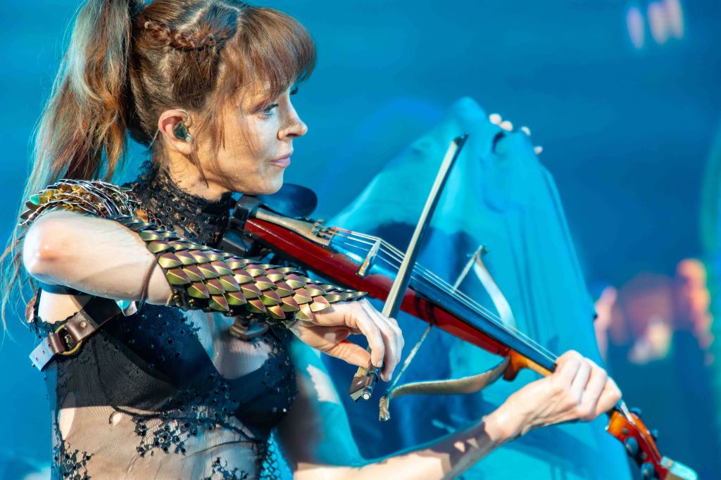 A musician playing the violin on stage, wearing a unique costume with metallic elements, against a colorful background.