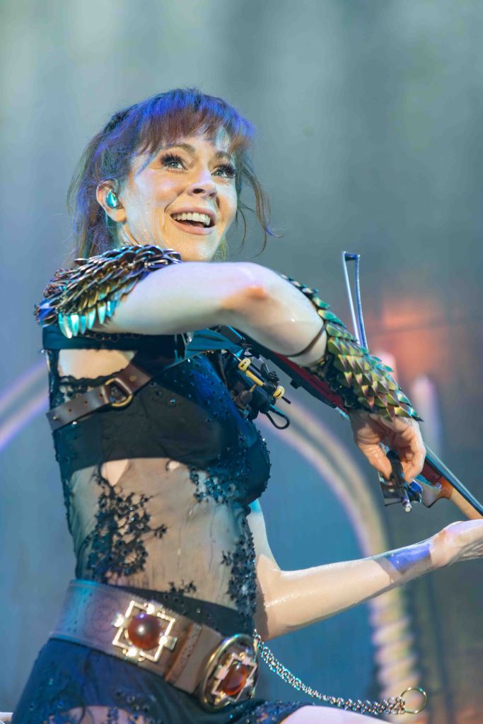 A female musician plays the violin on stage, smiling and wearing a stylish outfit with a lace design and embellished armor-like accessories, in a vibrant concert setting.