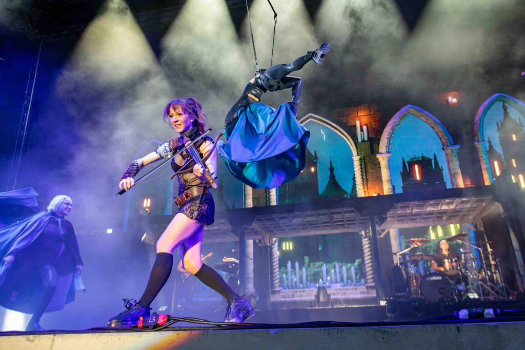 A performer on stage playing the violin while dancing, with an acrobatic figure suspended above them. The background features colorful lights and a gothic-inspired set. Another performer in a cloak can be seen in the shadows.