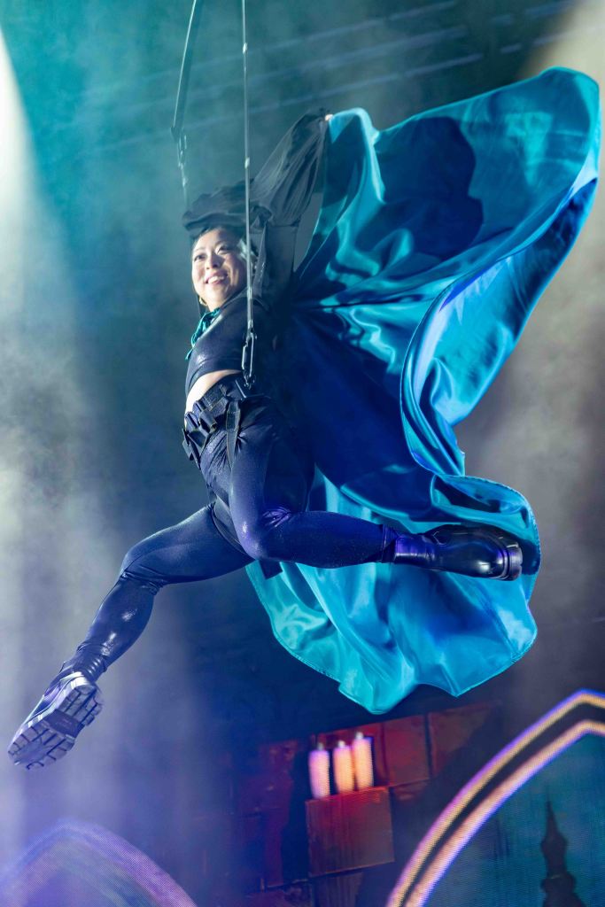 A performer in a black costume and flowing blue cape is suspended in mid-air, smiling while executing a dynamic leap on stage, surrounded by dramatic lighting and a smoky atmosphere.