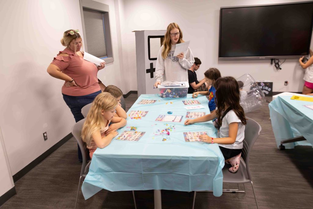 A group of children engaging in a game activity at a table covered with a light blue tablecloth, while an adult observes nearby; some kids are focused on game materials.