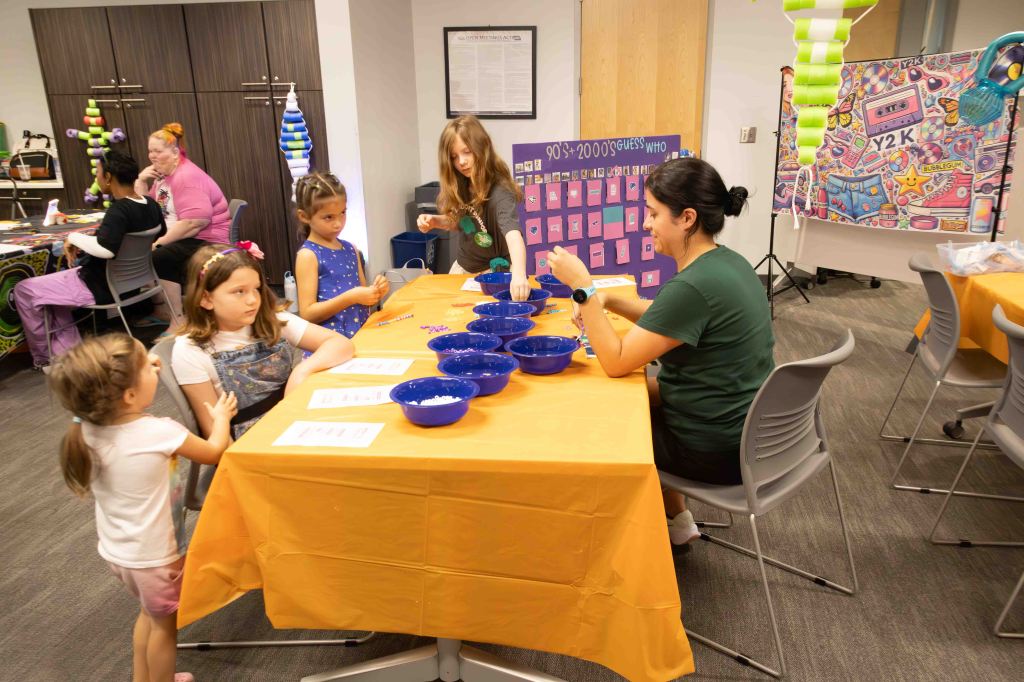 A group of children and adults engaged in a crafting activity at a table with orange tablecloths, featuring blue bowls and crafting materials. In the background, decorations and a display board related to the 90s and 2000s theme are visible.