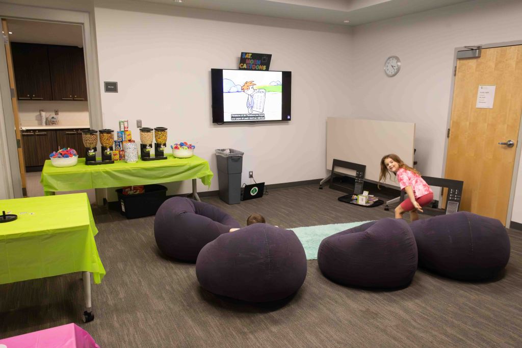 A colorful room with green tables, cozy bean bag chairs, and a television displaying a cartoon. There are snack dispensers on the table and two children interacting; one is seated on the floor and the other is standing nearby.