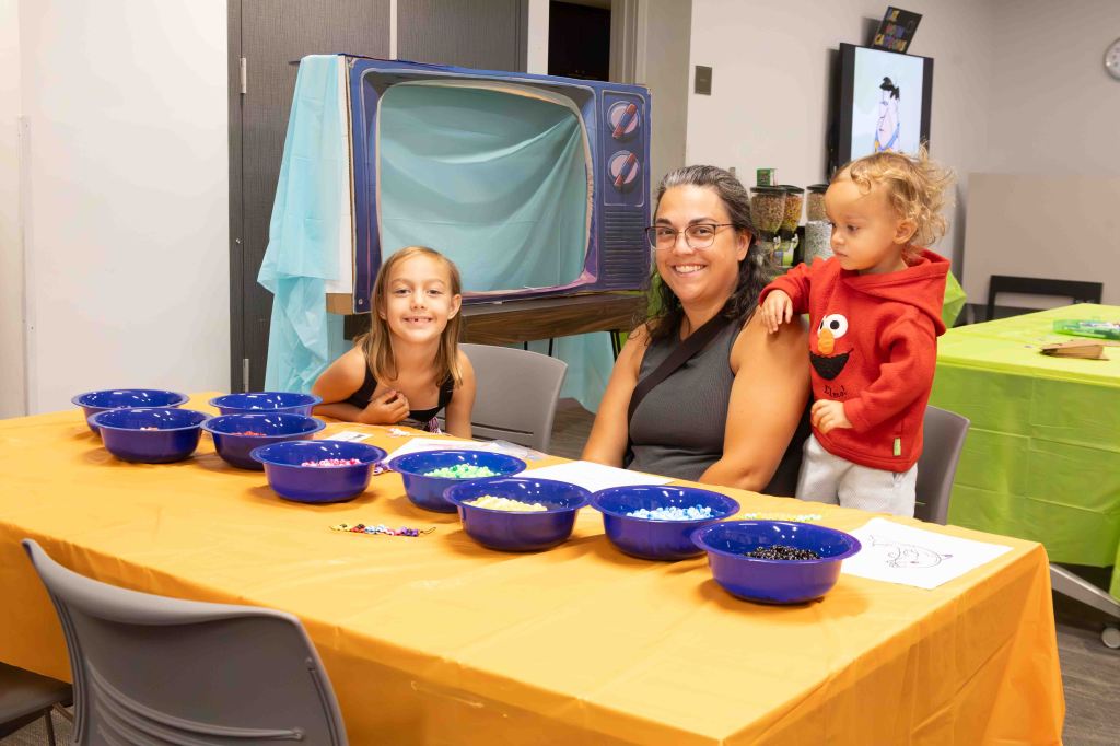 A woman sitting at a table with two children, surrounded by bowls of colorful beads, in a playful indoor setting.