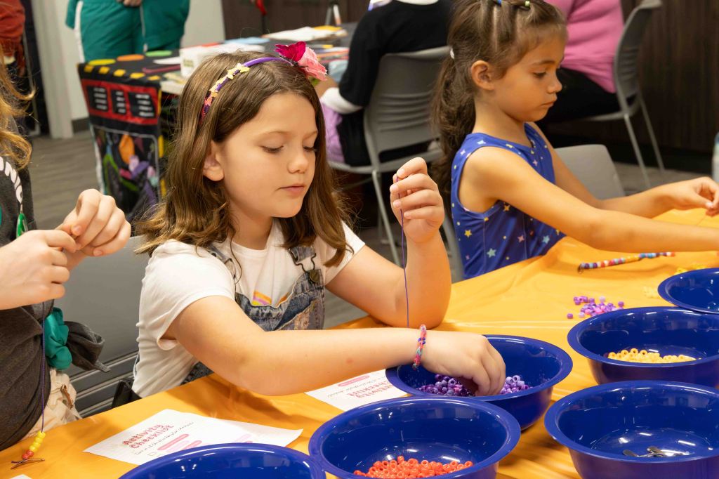 Two girls engaged in a beading activity at a table with blue bowls filled with colorful beads, focused on creating their bracelets.