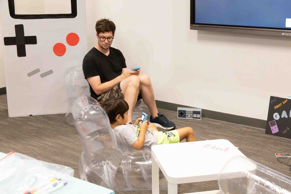 A man and a child sitting in transparent chairs, both looking at their devices, in a playful gaming-themed space with a large TV and game console decorations.