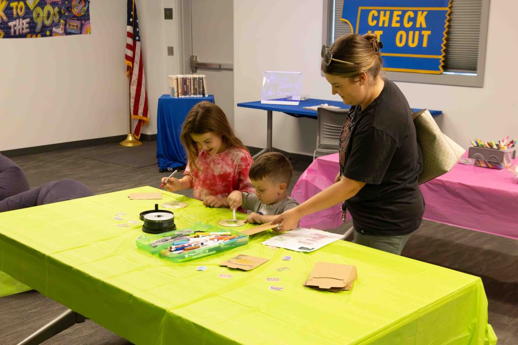 A woman and a girl engage in a craft activity at a bright green table, while a young boy looks on. The room has colorful decorations, including a banner and various markers and craft supplies scattered on the table.