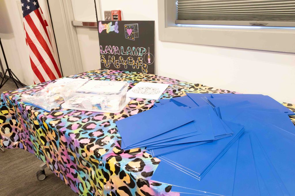 Table covered with a colorful leopard print cloth displaying craft supplies for a Lava Lamp Suncatcher activity, including blue construction paper and a clear container with materials.