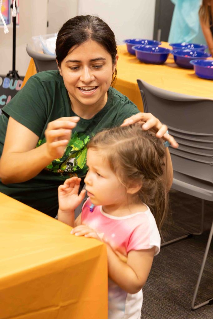 A woman with dark hair is engaged in a playful hairstyle activity with a young girl who looks curious. They are in a brightly lit room with orange tablecloths and blue bowls in the background.
