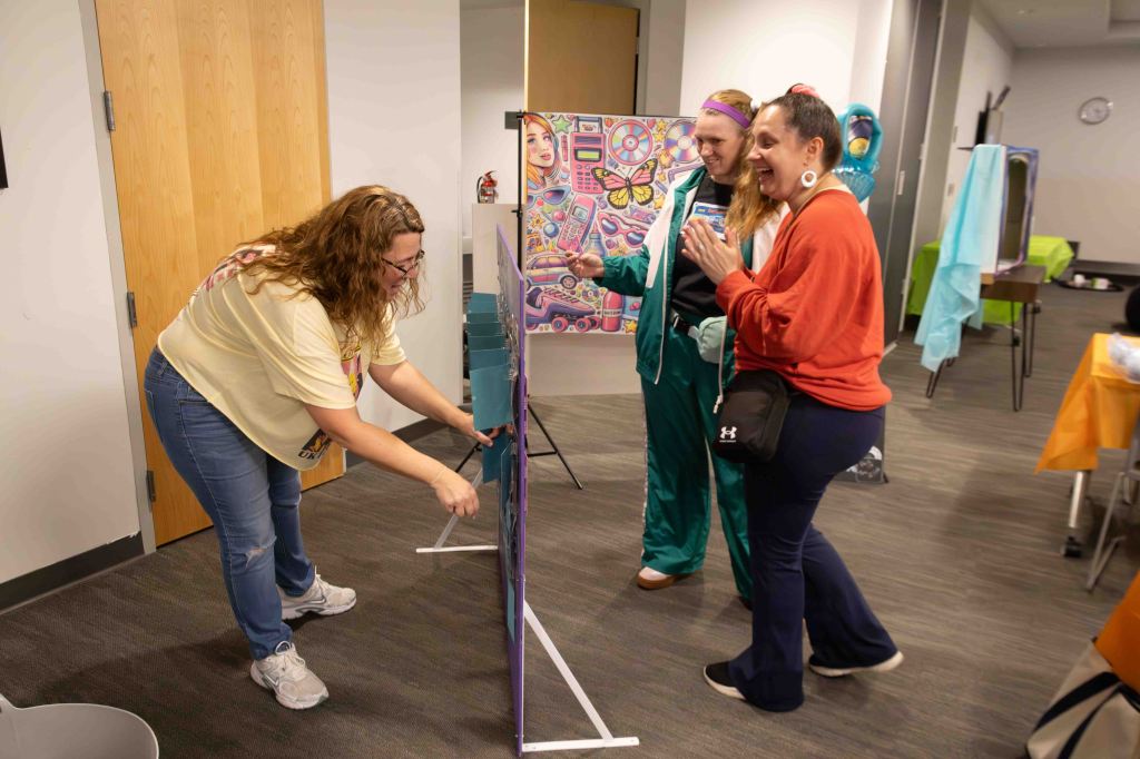Three women interacting in an indoor setting, with one woman adjusting a display board while the others observe and smile. Colorful artwork is visible in the background.