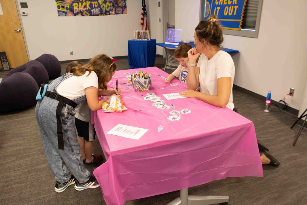 Children and an adult engaged in a crafting activity at a table covered with a pink tablecloth, surrounded by colorful markers and paper cutouts in a playful indoor setting.