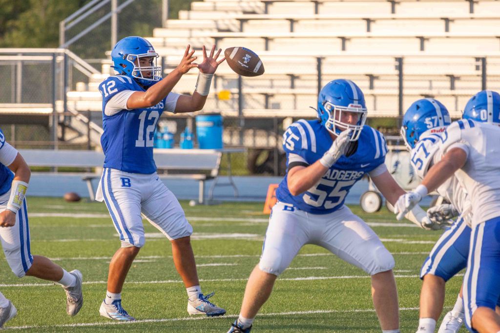 A high school football player wearing a blue jersey with the number 12 prepares to throw a football, while another player in a blue jersey with the number 55 blocks. The players are on a green football field with bleachers in the background.