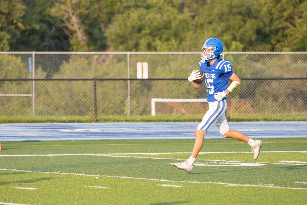A high school football player wearing a blue jersey with the number 15 runs on a grassy field during practice.
