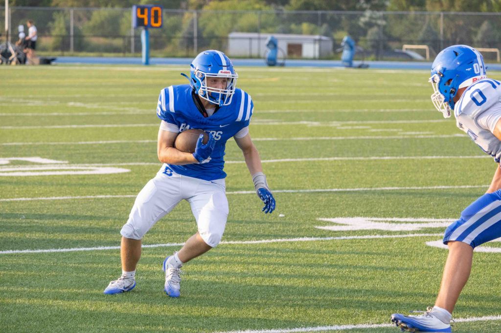 Football player in blue uniform running with the ball on a green field during a game.