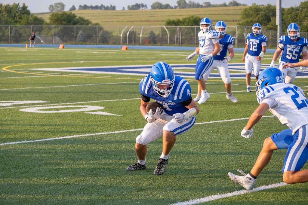 A football player in a blue and white uniform is running with the ball while being pursued by another player on a grassy field. More players in the background are positioned on the field.