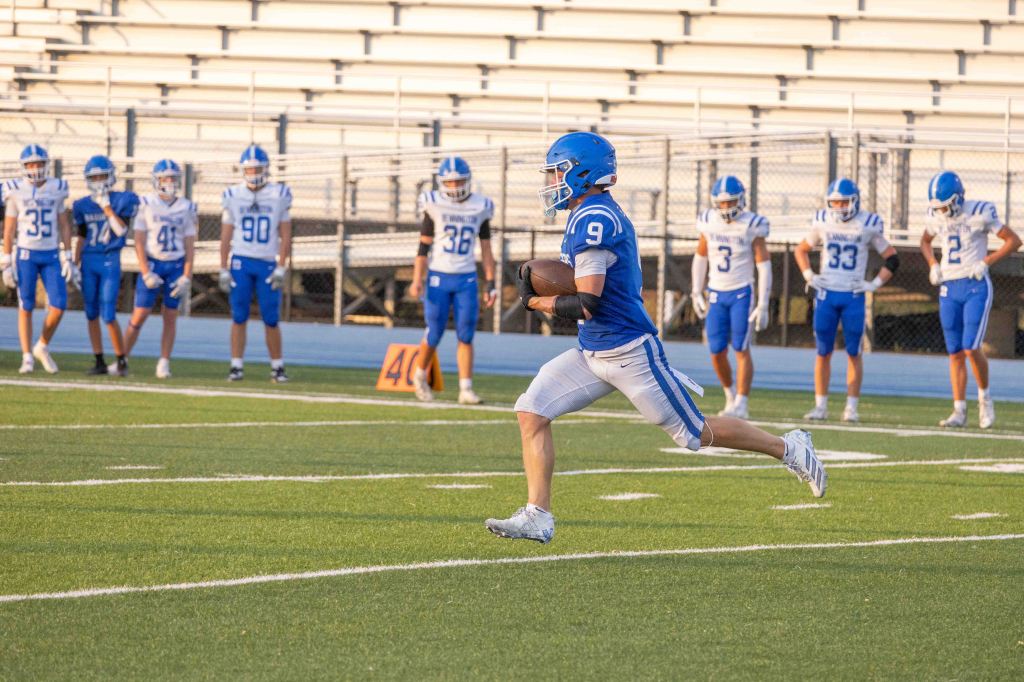 A football player wearing a blue jersey with the number 9 runs on the field, while teammates in white jerseys watch in the background.