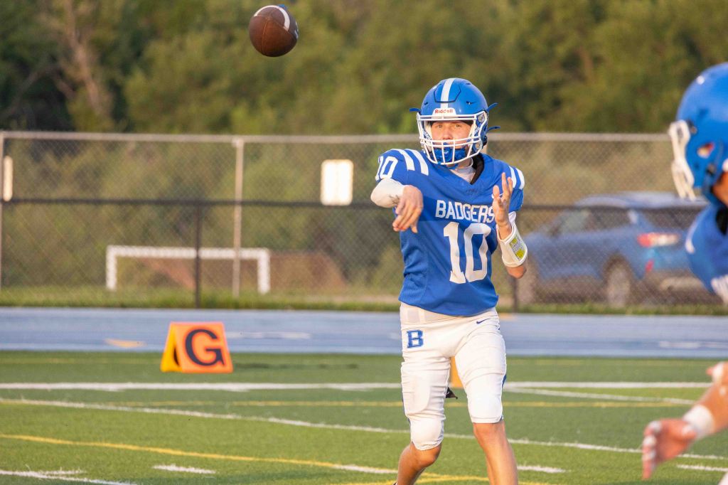 A young male football player in a blue jersey with 'Badgers' written on it throws a football on a field during a game.