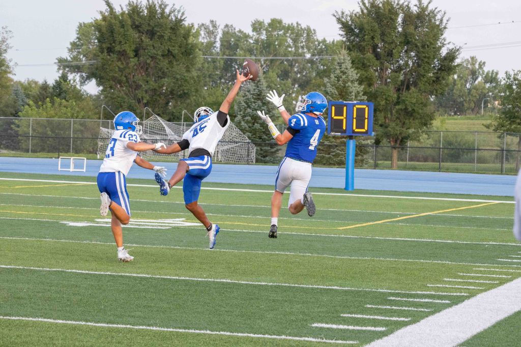 A football player in blue attempts to catch a pass while two defenders in white try to block him during a game on a grassy field.