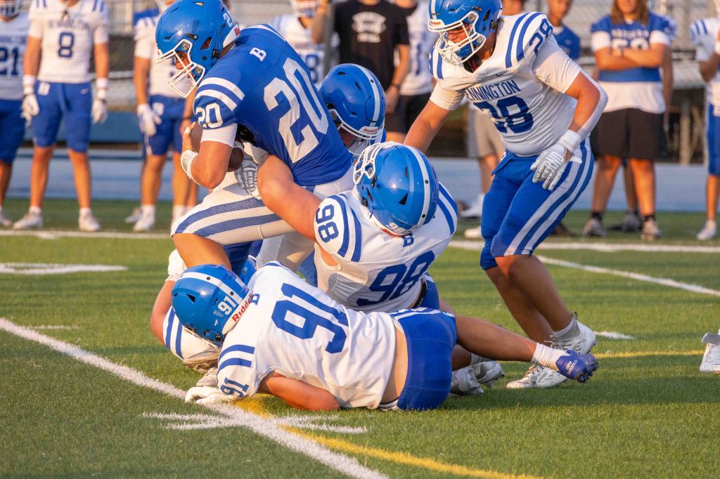 A football player in blue and white gear is tackled by multiple opponents on the field, with additional players in the background observing the play.