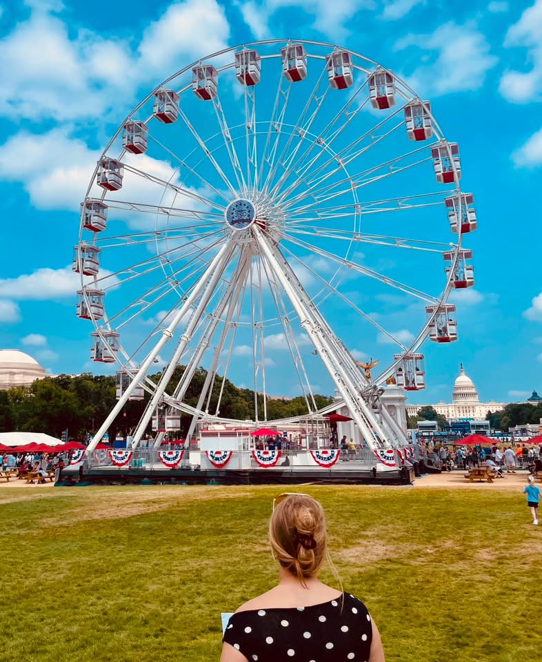 A woman with brown hair wearing a black polka-dot top stands in front of a large Ferris wheel at a festival, with the U.S. Capitol building in the background and a clear blue sky.