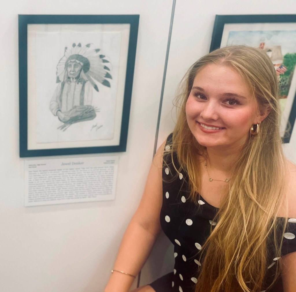 A young woman with long blonde hair and a polka dot dress is smiling next to a framed artwork of a Native American chief. The artwork is displayed on a wall, accompanied by a descriptive plaque.