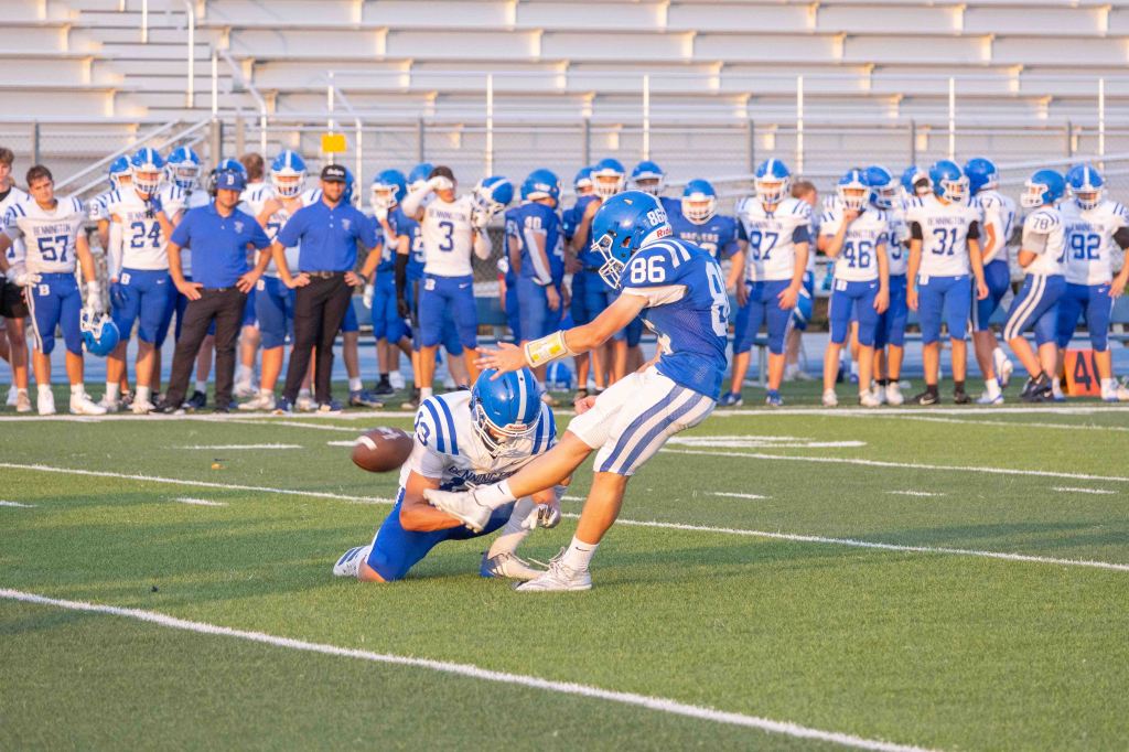 A football player prepares to kick a ball while another player kneels to hold it, with teammates in blue jerseys watching from the sidelines.