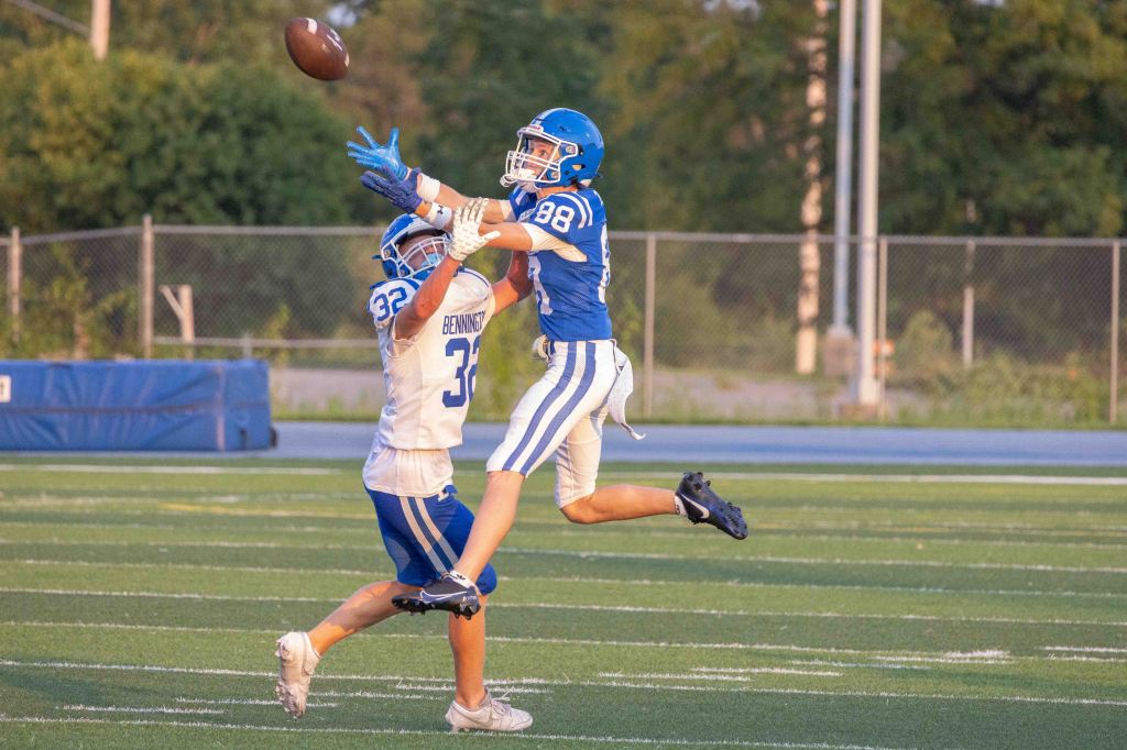 A football player in a blue jersey jumps to catch a flying ball while another player in a white jersey attempts to defend.