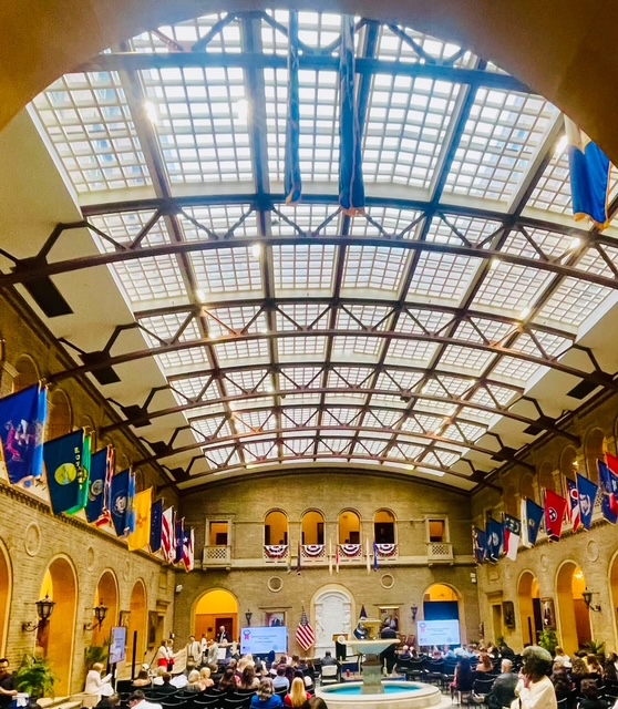 Interior view of a spacious hall with a vaulted glass ceiling, featuring numerous flags hanging from the walls. The room is filled with an audience seated around a central fountain, with a podium set for a speaking event.
