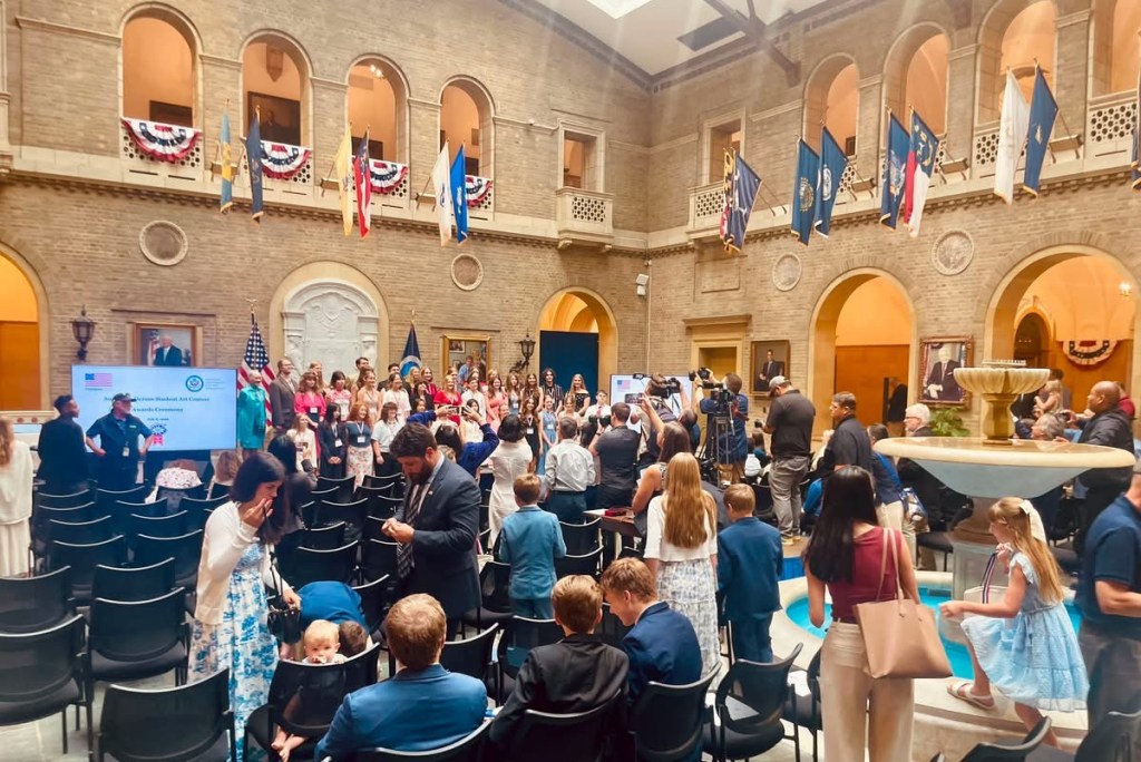 A gathering of children and adults in a spacious hall with a fountain, flags, and a presentation screen, during a ceremony.