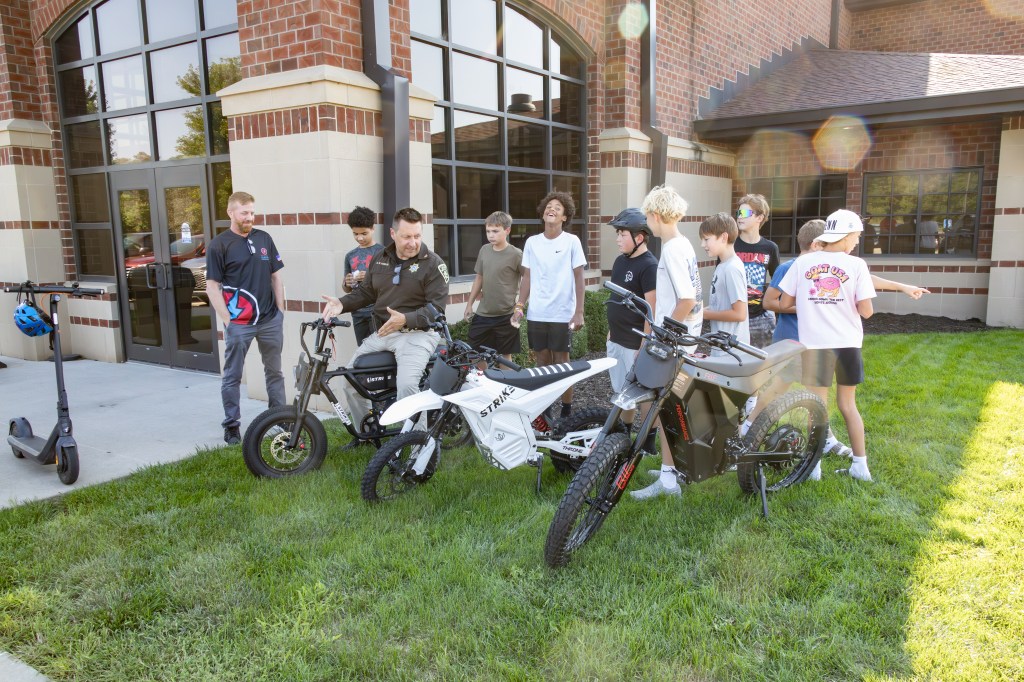 A group of children interacting with two electric bikes and a scooter outside a building, with adults nearby providing guidance.