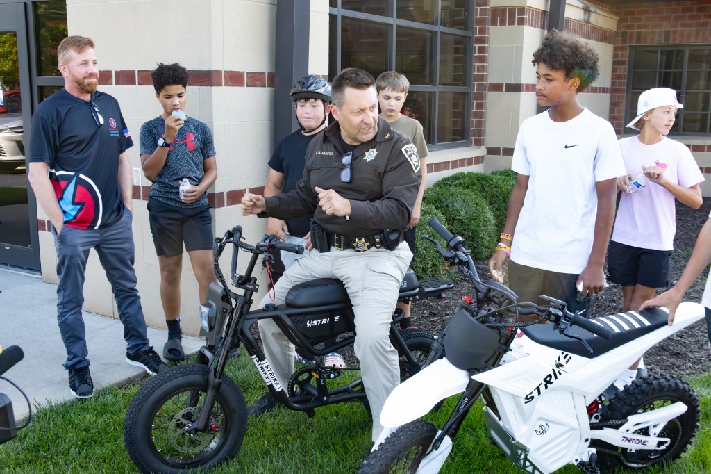 A police officer engages with a group of young people outside a building, with two electric bikes parked nearby. The officer is seated on a bike, while the youths listen and watch attentively.