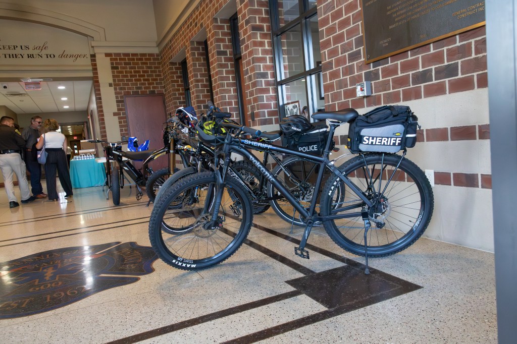 A row of police and sheriff bicycles parked in a lobby, with officers and guests engaged in conversation in the background.