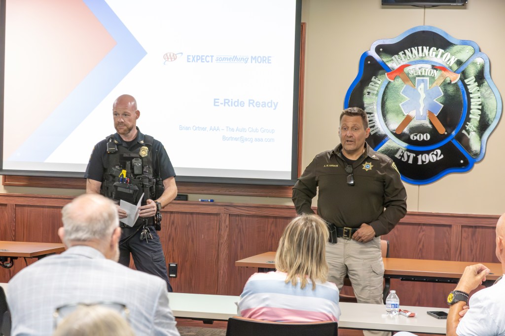 Two law enforcement officers presenting during a public safety event, with attendees seated in the foreground. A projector screen displays the title 'E-Ride Ready'.