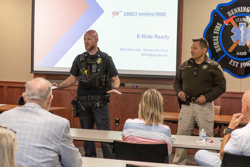 Two police officers speaking in front of an audience in a conference room during a presentation about E-Ride Ready, with a projector screen displaying information.