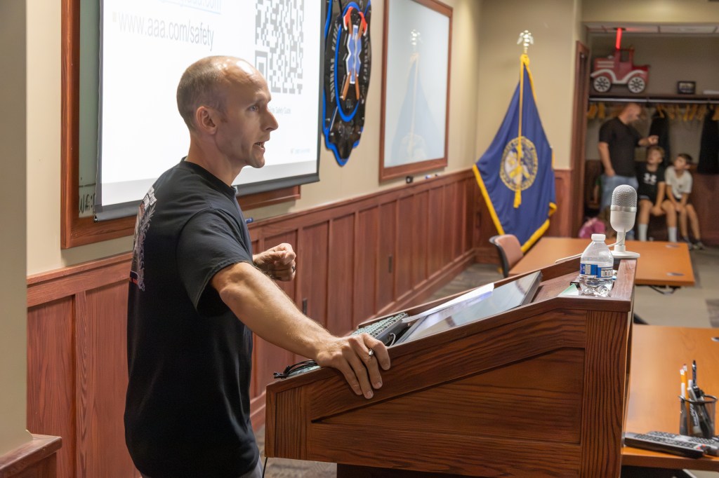 A speaker engages an audience from a podium in a classroom or conference setting, with a presentation on a screen behind them and a flag displayed nearby.