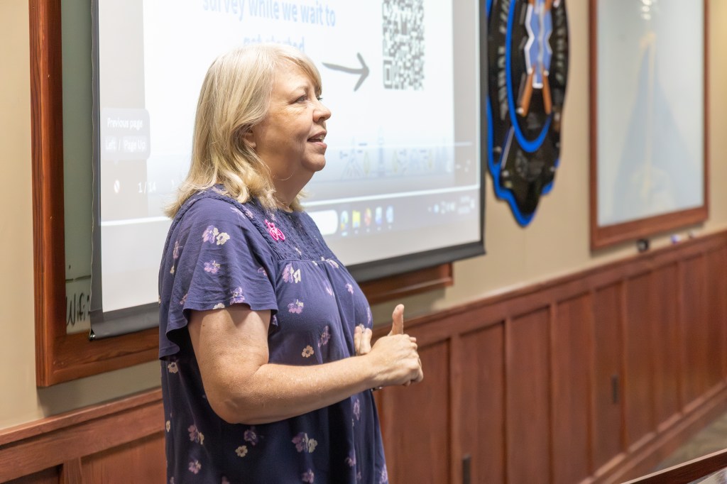 A woman standing in front of a presentation screen, engaging an audience with her hands raised in a casual gesture.
