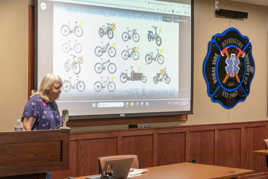 A woman stands by a podium in a room, presenting in front of a large screen displaying various illustrations of bicycles with symbols showing different statuses. An emblem of a fire district is visible on the wall.