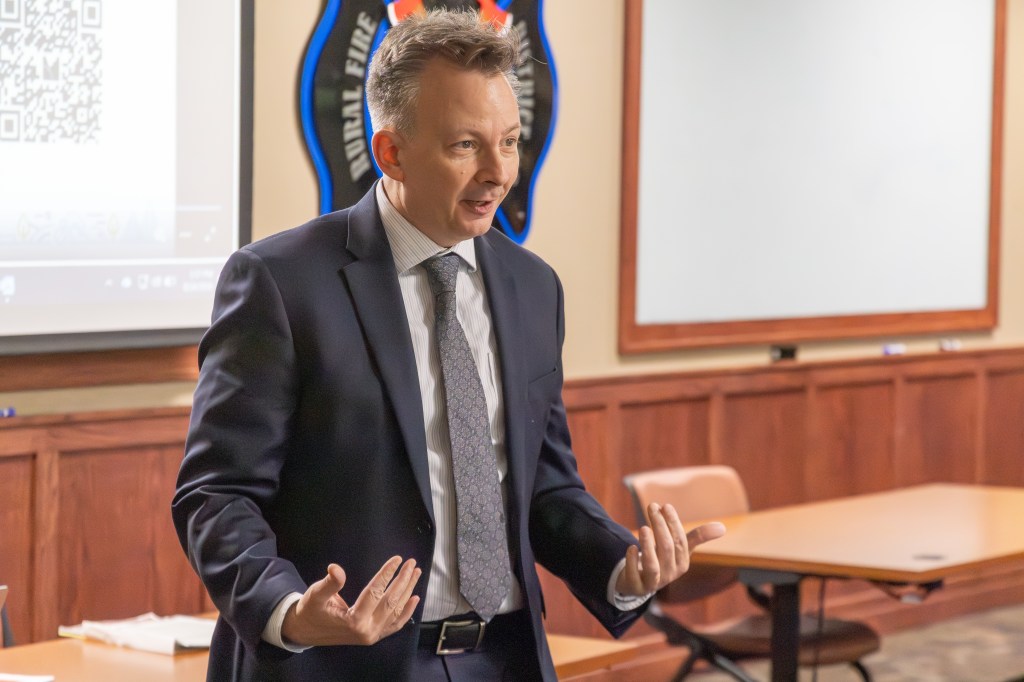 A business professional in a suit presents to an audience in a conference room, gesturing with his hands while speaking.