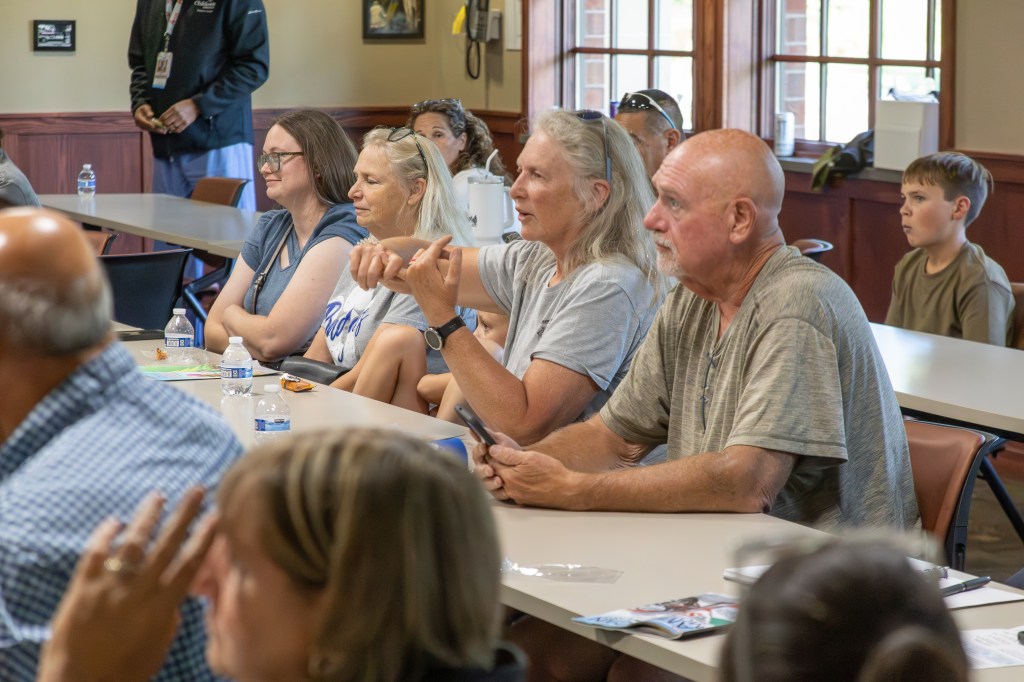 A group of people sitting at tables in a meeting or presentation, showing engaged expressions. Multiple adults are seated, with a focus on one man and one woman who appear to be discussing or asking questions.