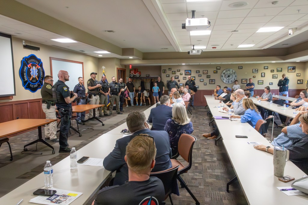 A group of law enforcement officers stands in front of an audience during a community meeting, with tables set up in a conference room. Various attendees are seated, engaging with the presentation.