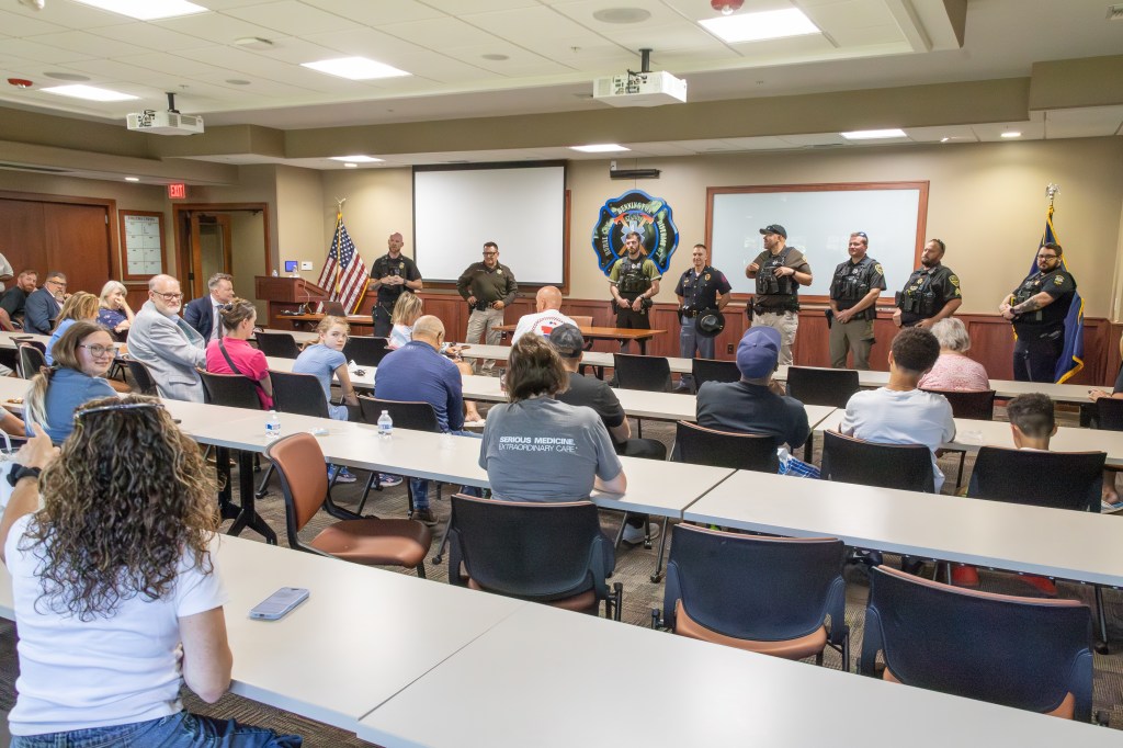 A group of law enforcement officers standing in front of an audience in a meeting room, with tables arranged for attendees. The officers are facing the crowd, who are seated and engaged in the presentation.