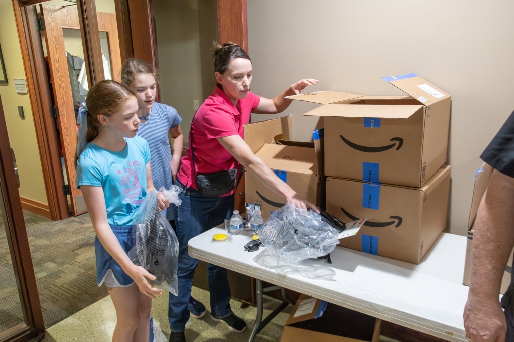 A woman and two girls unpack items from large Amazon boxes on a table in a hallway.