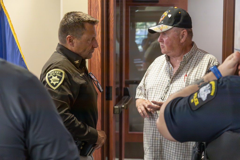 A sheriff in a dark uniform engages in conversation with an older man wearing a checkered shirt and a hat. In the background, other police officers are partially visible.