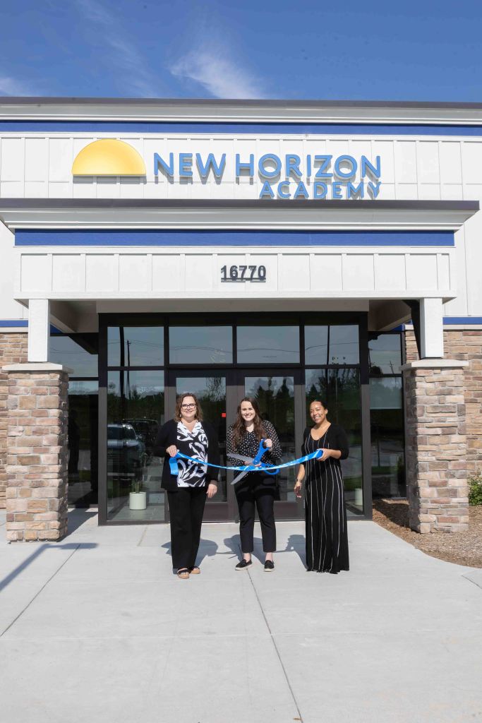 Three women holding a large blue ribbon in front of the New Horizon Academy building during a ribbon-cutting ceremony.