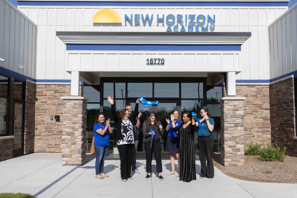 A group of individuals celebrating outside the New Horizon Academy building, with a ribbon and scissors, during a ribbon-cutting ceremony.