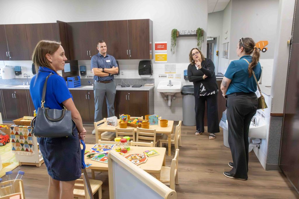 A group of four adults engages in discussion in a classroom setting designed for young children, featuring play areas and educational toys.