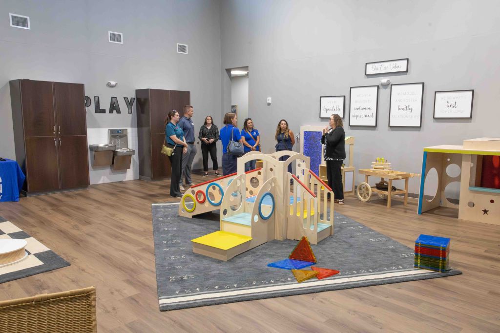 A group of adults observes a colorful play area featuring wooden play structures, toys, and educational materials, with motivational quotes displayed on the walls.