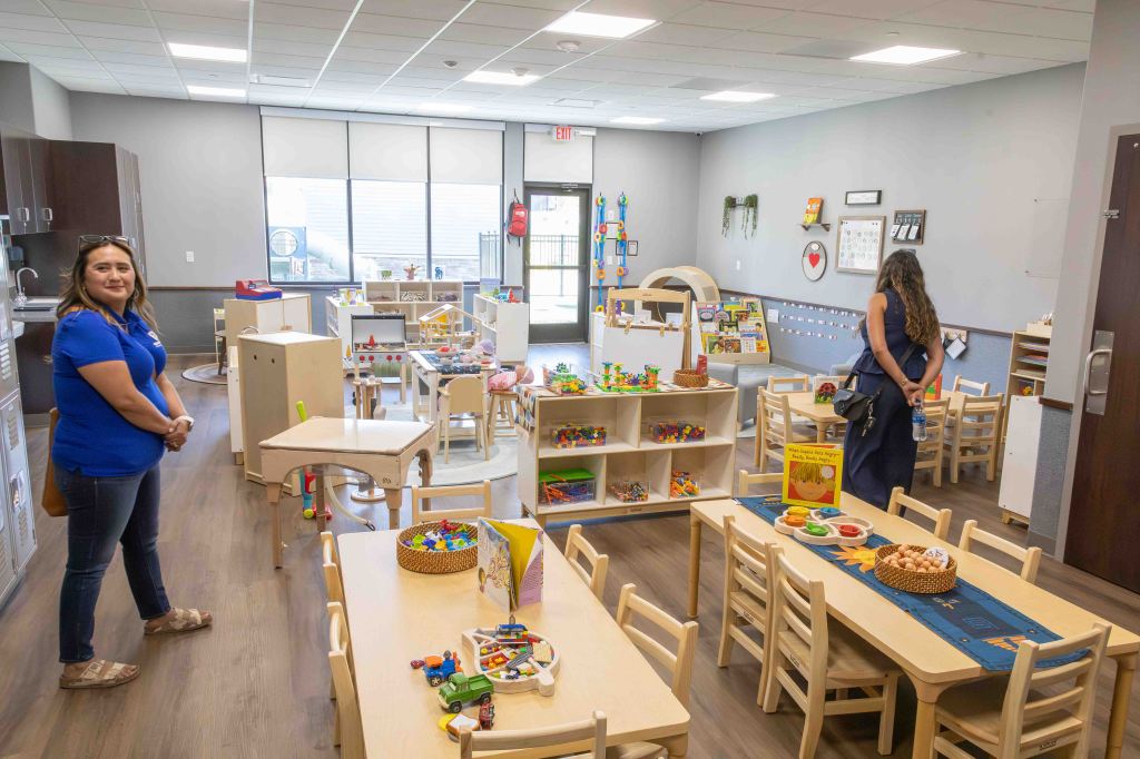 A bright and colorful classroom filled with various toys and learning materials. Two women are present, one standing near the entrance wearing a blue shirt and the other observing from a distance. The room features wooden furniture, activity tables, and shelves filled with educational toys.