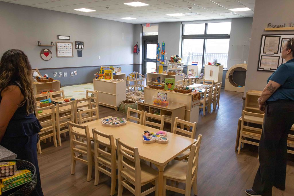 A brightly lit preschool classroom featuring wooden tables and chairs, decorated with colorful educational materials and toys. Two adults are present, engaging in conversation, while various play areas are visible in the background.