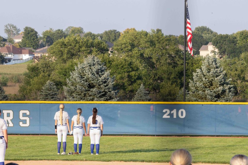 Four female softball players standing with their backs to the camera, facing a flagpole with an American flag, in a grassy field with trees and houses in the background.